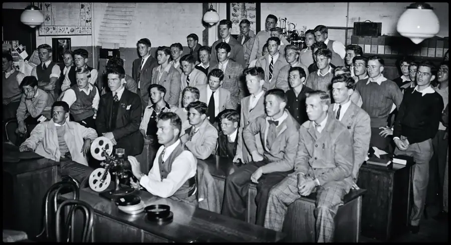 A group of young men in suits sits and stands in a classroom, watching a film projector operated by one person. Vintage lights hang from the ceiling, and bookshelves line the walls.