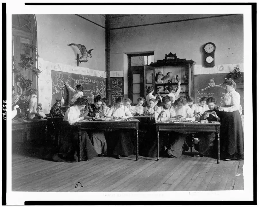 Black-and-white photo of girls in a classroom, drawing birds at their desks. A teacher stands nearby. Chalkboards, bird models, and a clock decorate the room.