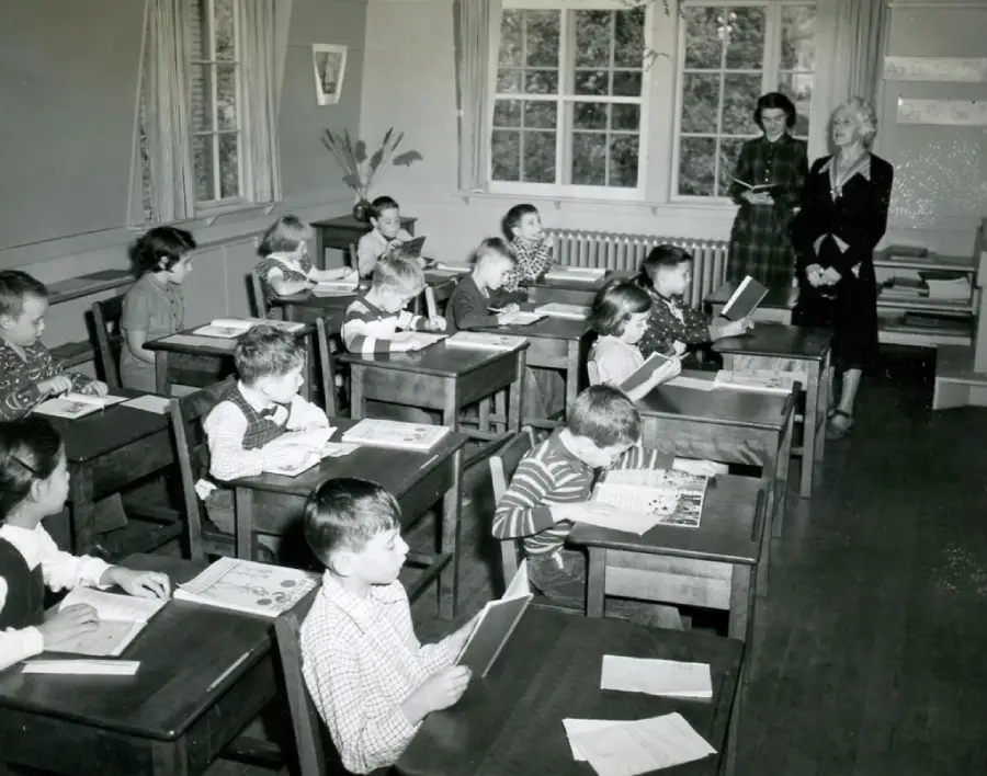 Black-and-white photo of young children sitting at desks in a classroom, reading books. Two women stand at the front near a window, possibly teachers. The classroom has wooden desks and large windows letting in light.