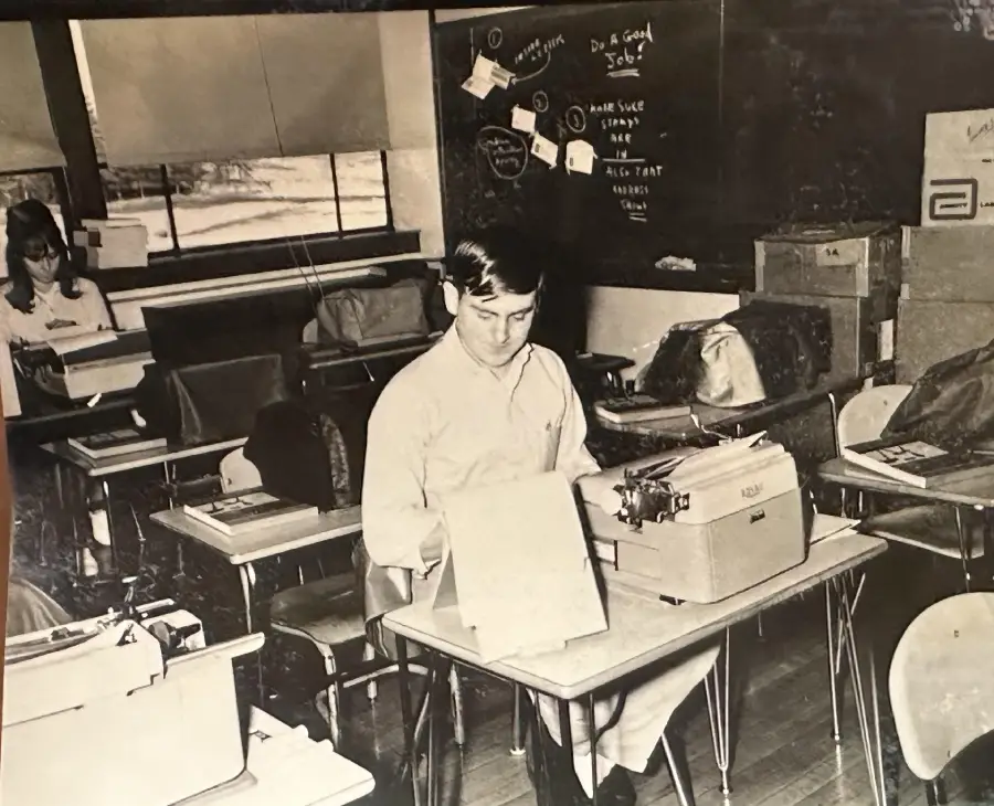 A man sits at a desk using a typewriter in a classroom with other desks, papers, and boxes. The chalkboard behind him has notes and messages written on it. Another person sits in the background by the window.