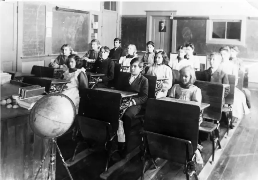 Black and white photo of children sitting at old-fashioned desks in a classroom. A globe is visible in the foreground, and chalkboards cover the walls behind the students. The children face forward, looking at the camera.