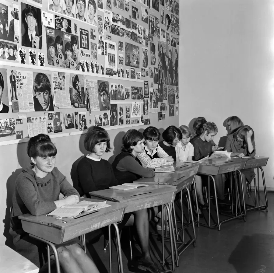 A row of girls sit at desks in a classroom with walls covered in Beatles posters and magazine clippings.