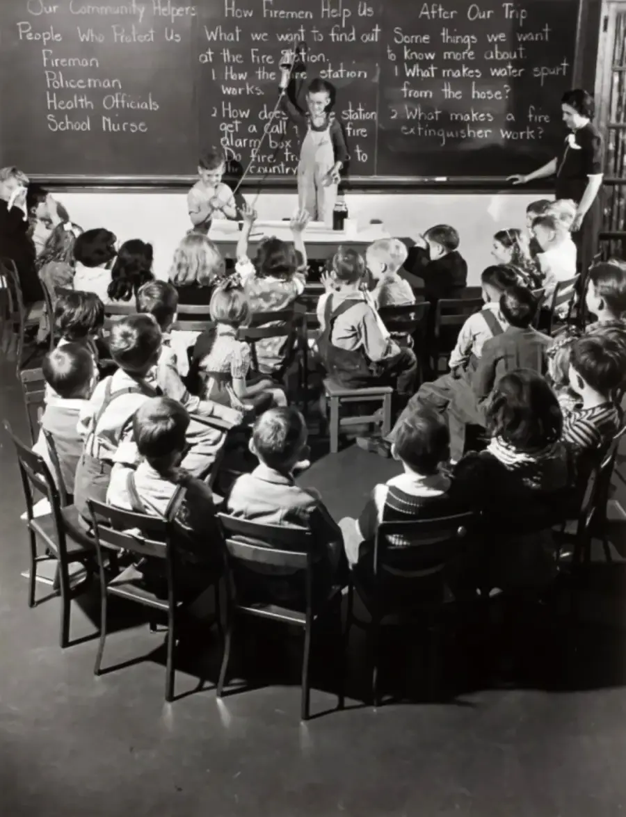 Black and white photo of a classroom. Children sit facing a teacher and two adults at the front. Lessons on "firemen" and safety are written on the chalkboard behind them.