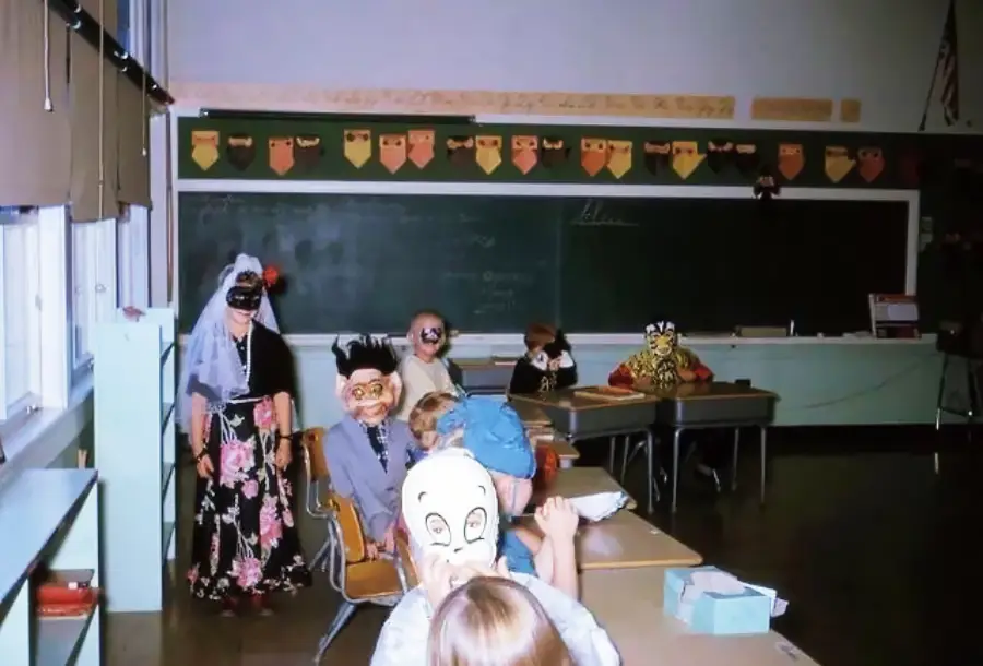 Children in Halloween costumes and masks pose in a classroom with desks, a chalkboard, and decorations on the wall.