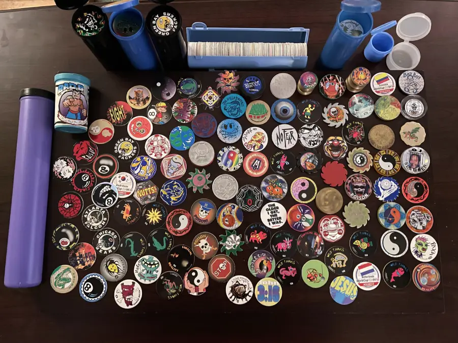 A collection of colorful POGs and Slammers with various designs spread on a table, surrounded by containers and tubes used for storing and playing with them.
