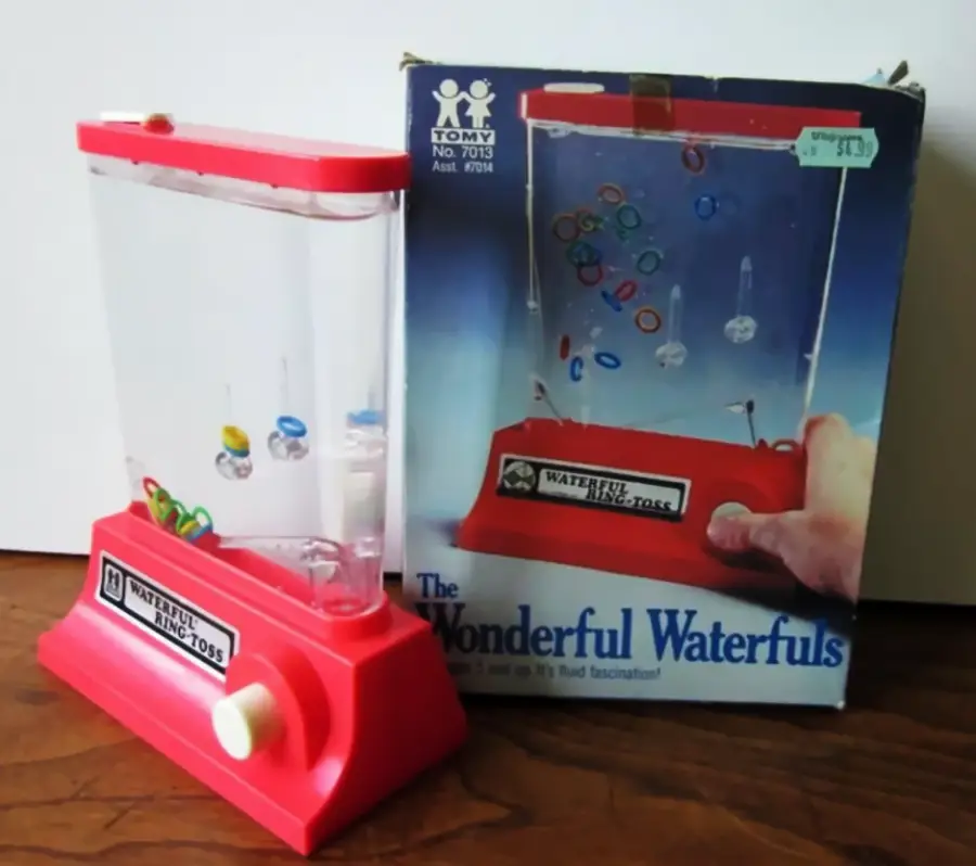 A vintage Tomy Wonderful Waterfuls handheld water game sits on a wooden surface next to its original box, featuring colorful rings and a white button for gameplay.