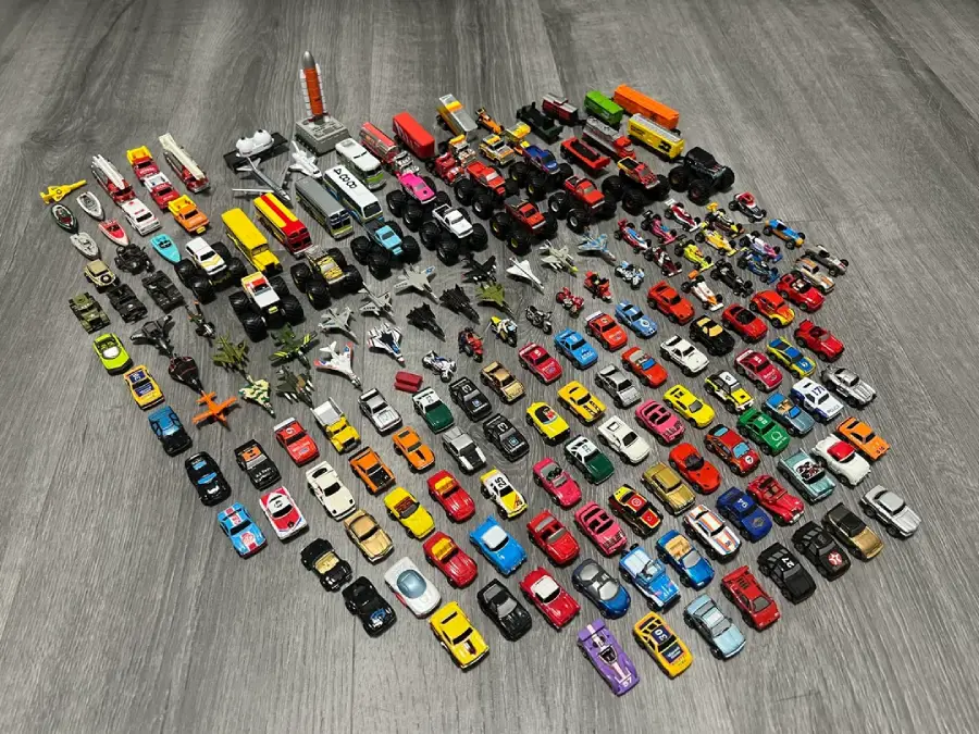 A collection of toy vehicles, including cars, trucks, planes, boats, and helicopters, is neatly arranged in rows on a wooden floor.