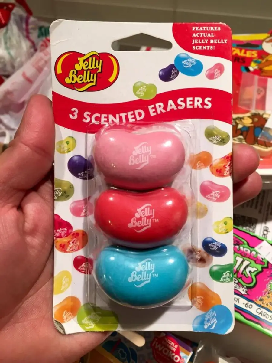 A hand holds a pack of three Jelly Belly scented erasers shaped like jelly beans in pink, red, and blue, with colorful jelly bean graphics on the packaging.
