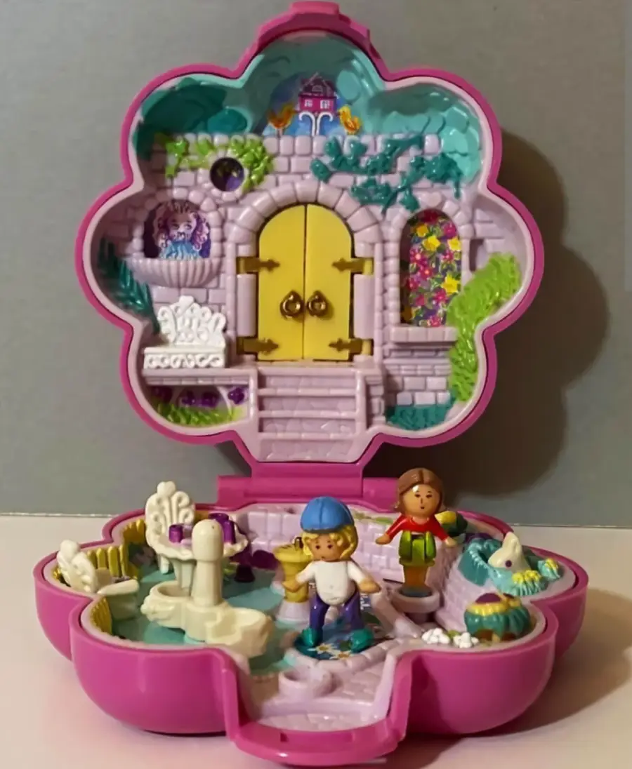 A colorful, flower-shaped toy case opens to reveal a miniature garden scene with two small figures, a white fountain, greenery, and a purple castle door in the background.