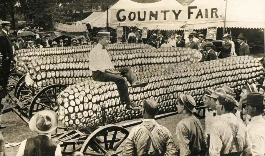 Black and white photo of a man sitting on a parade float shaped like large ears of corn, surrounded by people at a county fair, with a “County Fair” tent sign in the background.
