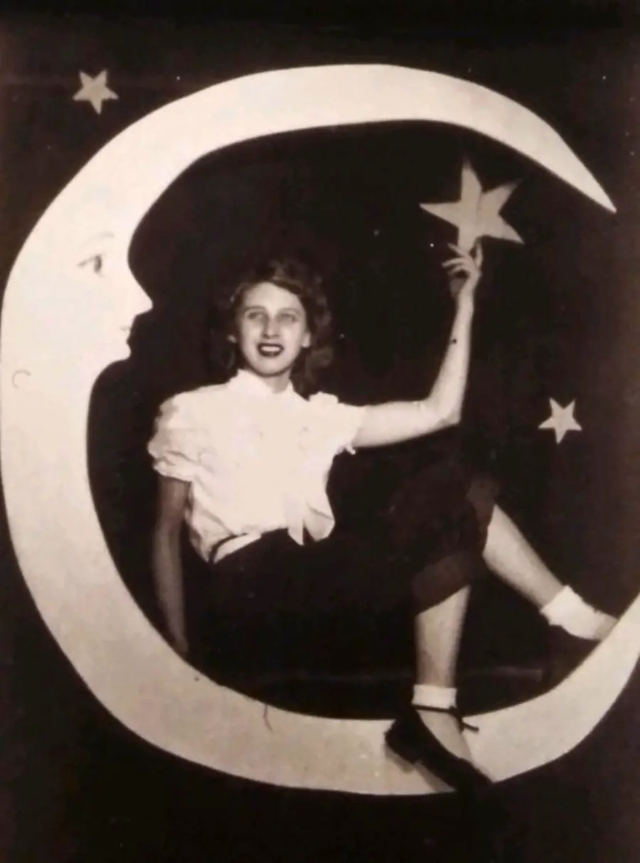 A woman sits on a crescent moon cutout, holding a star cutout, with stars and a face on the moon. The photo is black and white, giving it a vintage look.