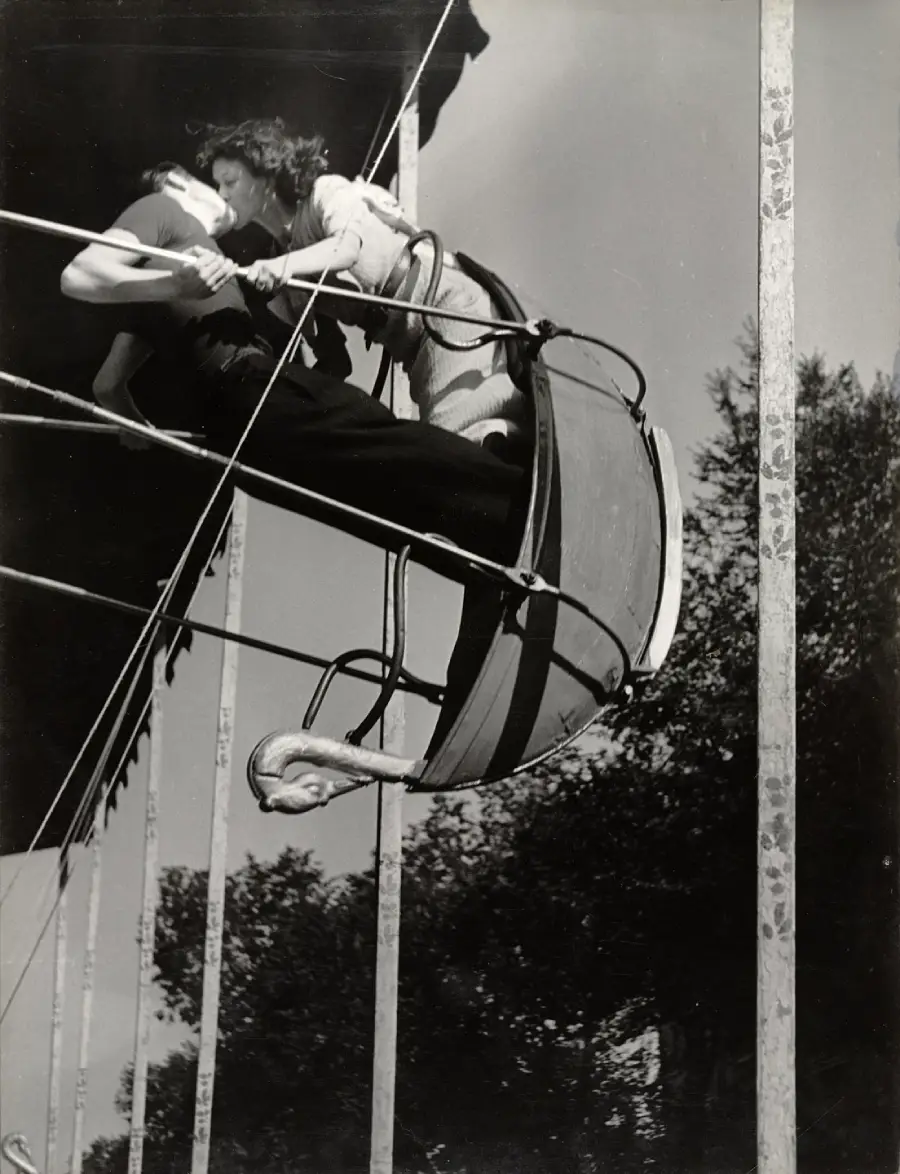 A couple shares a kiss while riding a swan-shaped swing ride at an amusement park, captured at the ride’s highest point against a clear sky and trees.
