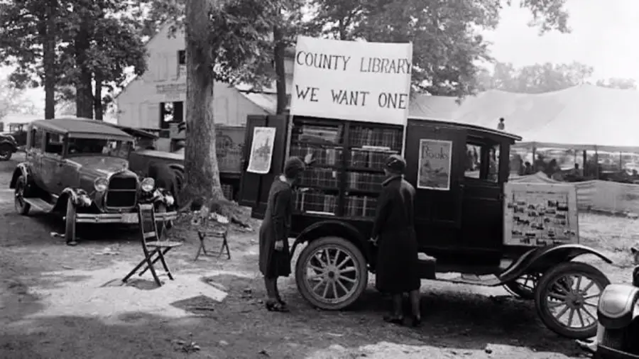 Two women stand by a vintage vehicle displaying books, with a sign reading "County Library—We Want One." Other cars, chairs, and trees are nearby, suggesting a rural outdoor event in the early 20th century.
