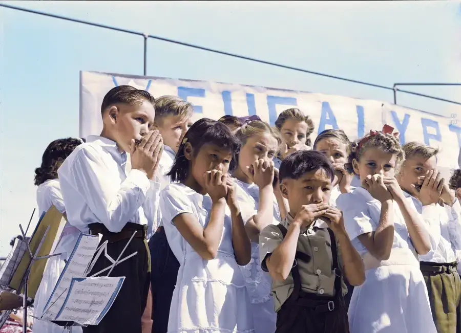 A group of children, dressed formally, stand together outdoors playing harmonicas in front of a partially visible sign. Some hold sheet music while performing.