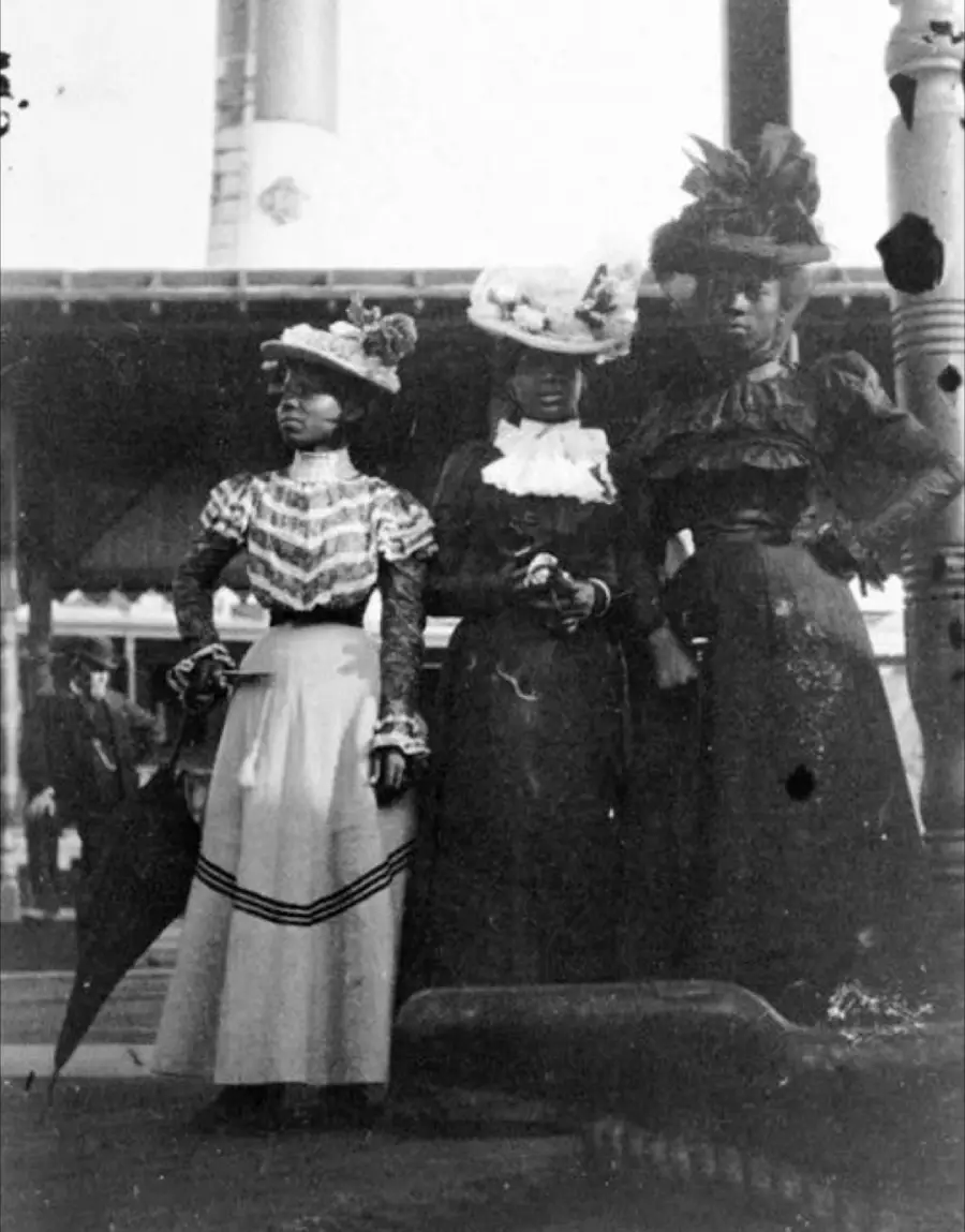 Three Black women in elegant late 1800s dresses and elaborate hats stand together outdoors, one holding a closed umbrella. The background includes a building and a tall smokestack.