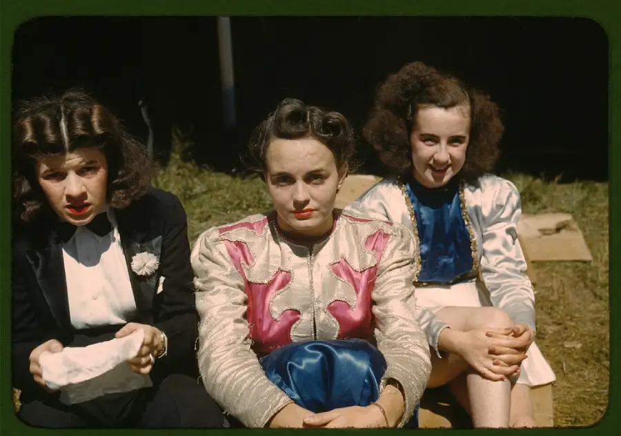Three young women sit on the grass, wearing vintage 1940s clothing. Two wear colorful jackets, one in a dark blazer. They look toward the camera, with expressions ranging from serious to slight smiles.
