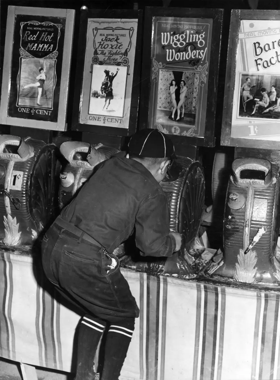 A boy peers into a vintage arcade machine at a penny arcade, with several themed peep show machines labeled “Red Hot Mama” and “Wiggling Wonders” lined up in the background.