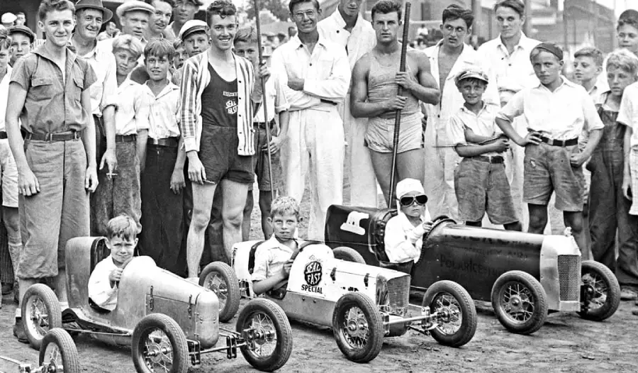 Three boys sit in small race cars at the starting line, surrounded by a crowd of boys and men, some in summer clothes, all outdoors on a dirt surface, giving a festive and competitive atmosphere.