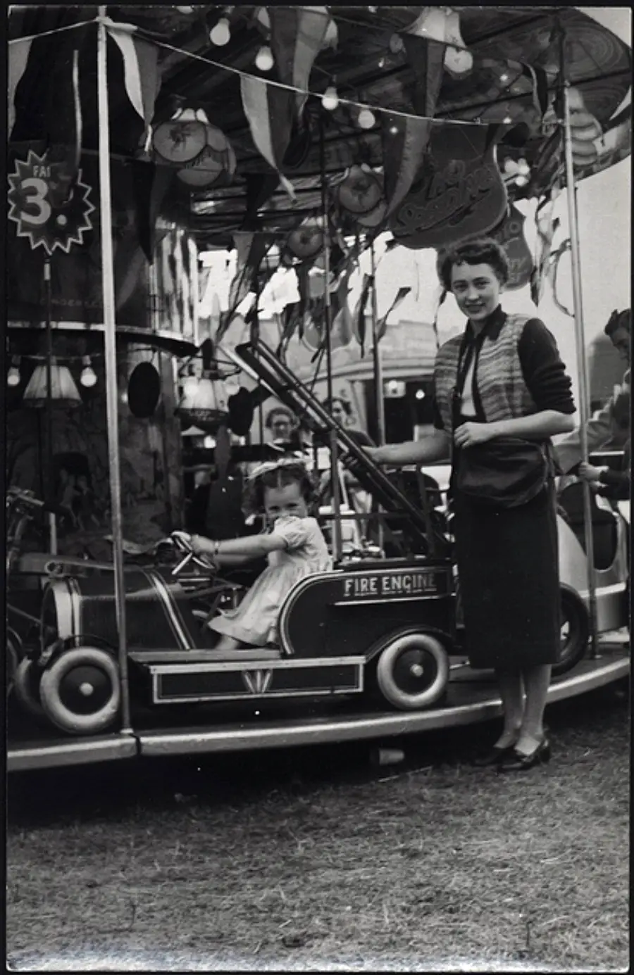 A woman stands by a merry-go-round with a smiling child seated in a fire engine ride. There are decorations, lights, and people in the background at what appears to be a fair.