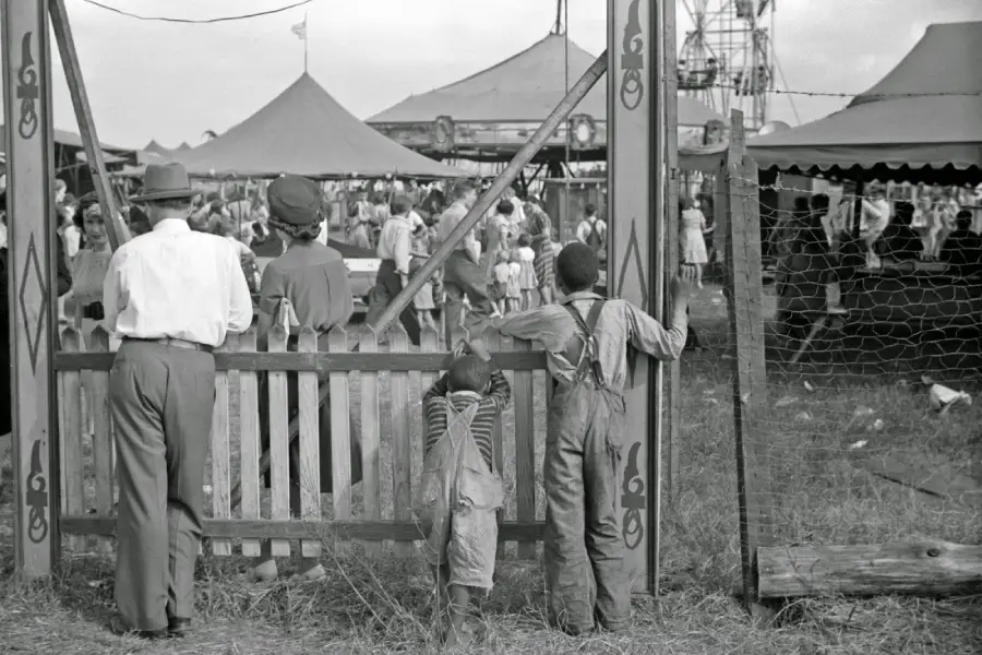 Three people stand at a wooden fence watching a busy carnival scene with tents, a Ferris wheel, and crowds in the background. Two boys wear overalls; one adult wears a hat and suit.