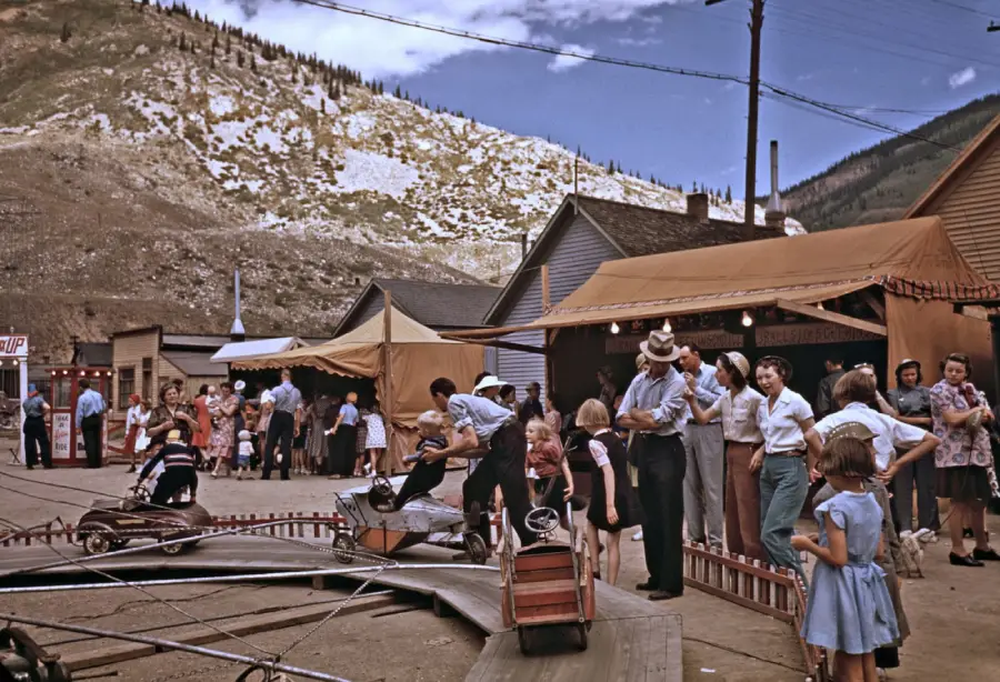 People gather at an outdoor fair in a mountain town, with booths, children riding small cars, and adults watching. Snowy hills and blue sky are in the background. The scene appears to be from the mid-20th century.