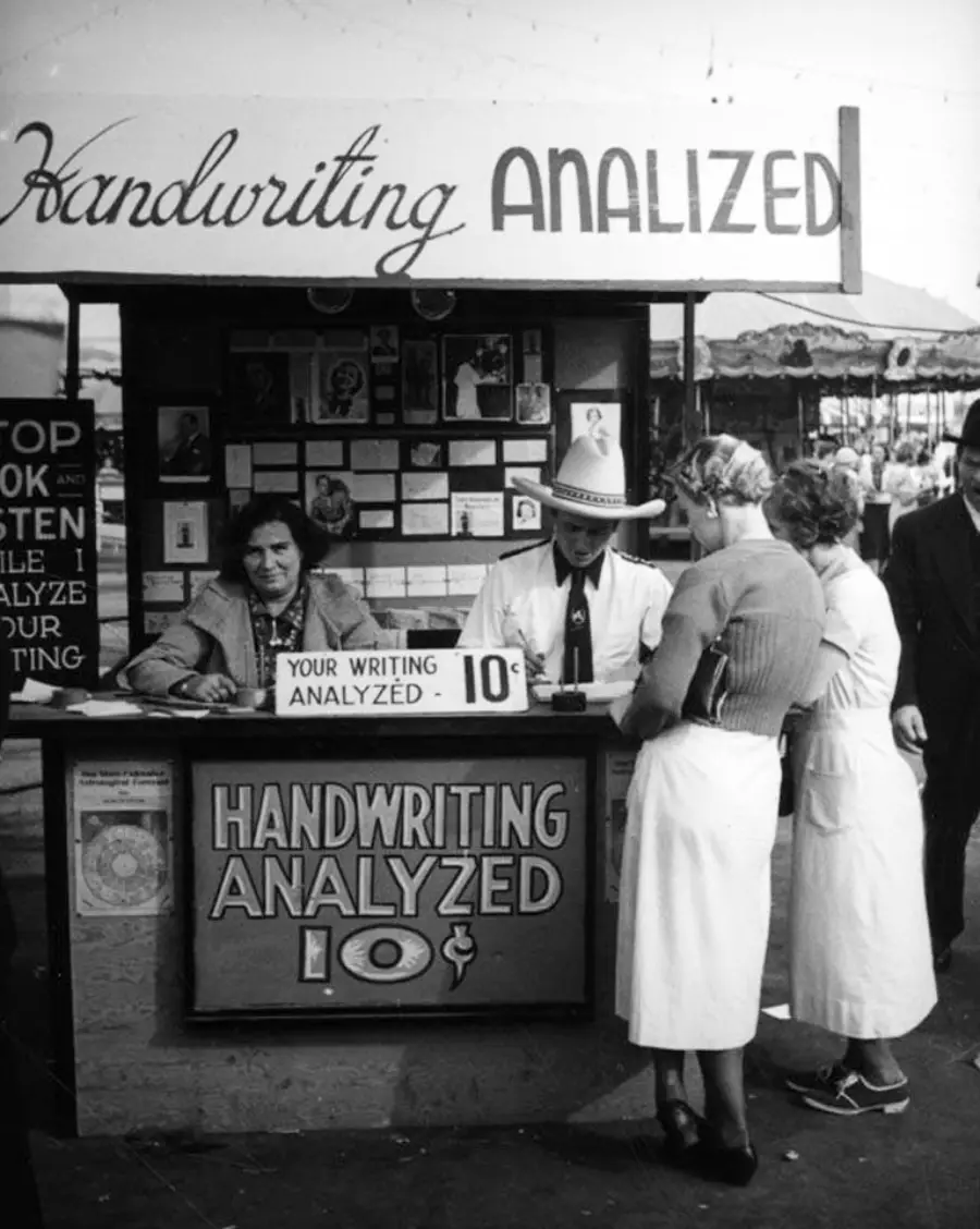 Black and white photo of a booth labeled "Handwriting Analyzed 10¢," with a man and woman analyzing handwriting for two customers at an outdoor market or fair. Signs and people are visible in the background.