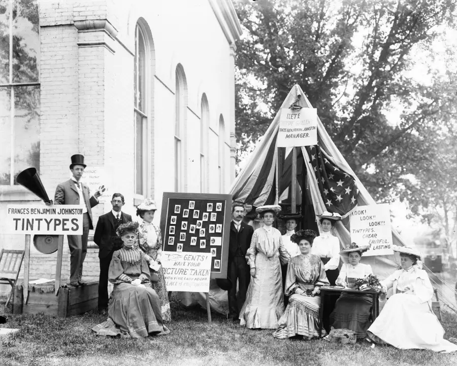 A group of men and women in vintage clothing pose outside a tent with signs advertising tintype photography. Some sit and some stand, with props including cameras and framed photos, in front of a brick building and trees.