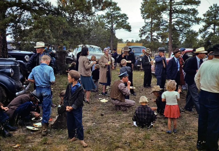 A group of people, including adults and children, gather outdoors near trees and vintage cars, eating and socializing at what appears to be a picnic or community event.