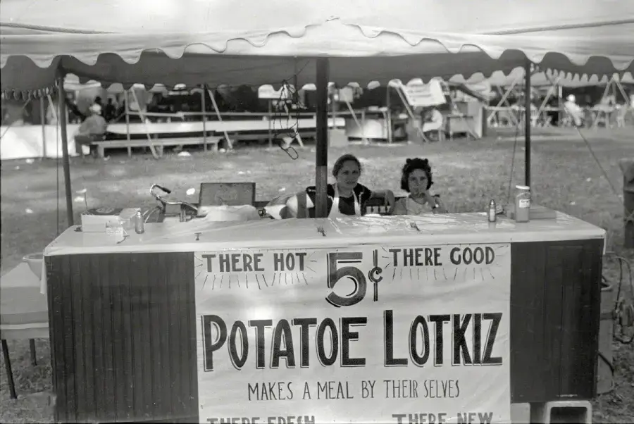 Two women sit behind a booth at an outdoor event, selling "Potatoe Lotkiz" for 5 cents. The sign reads "There hot, there good, makes a meal by their selves." Tables and people are visible in the background.