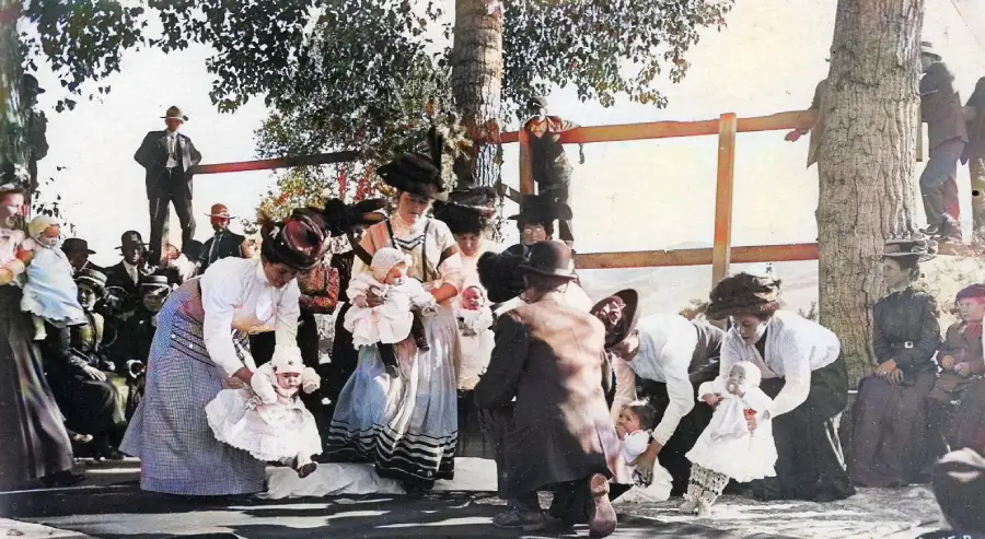 A group of women in vintage clothing hold babies outdoors near trees, while people watch and children kneel on the ground. Some spectators sit or stand in the background.