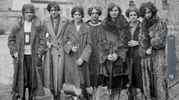 Seven women in vintage coats stand in a line outdoors, each holding a rifle. The image is black and white, suggesting it was taken in the early 20th century. The background shows blurred trees and a building.