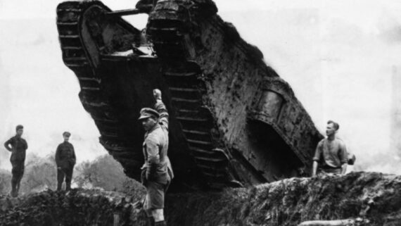 A large World War I tank crosses a trench while several soldiers stand nearby, one raising his arm, in a black-and-white photo.