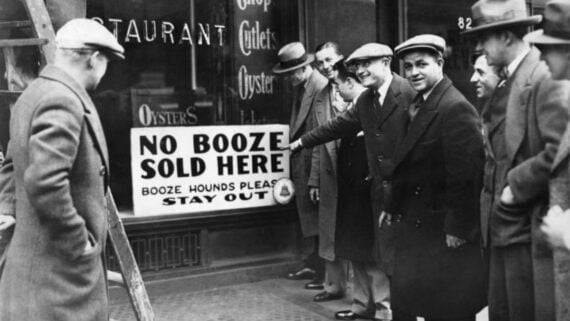 A group of men in coats and hats stand outside a restaurant. One man points to a sign in the window that reads, &ldquo;NO BOOZE SOLD HERE. BOOZE HOUNDS PLEASE STAY OUT.&rdquo;