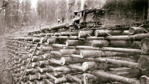 A steam train crosses a bridge made entirely of stacked logs in a forested area, with workers standing near the train on top of the logs.