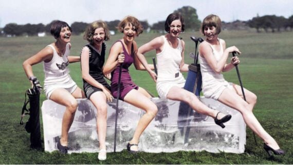 Five women in vintage swimsuits sit and laugh on a large block of ice outdoors, each holding a golf club, with a grassy field and trees in the background.