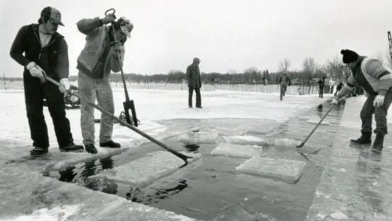 Four people cut and move large blocks of ice from a frozen lake using long tools. Snow covers the ground, and more people can be seen in the wintry background. The scene appears cold and overcast.