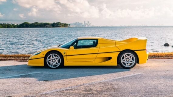 A yellow Ferrari sports car parked by the water, with a city skyline and clouds visible in the background.