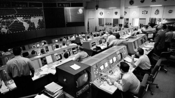 Black-and-white photo of a busy NASA mission control room with rows of engineers at computer consoles, large screens displaying world maps, and various technical equipment and displays on the walls.