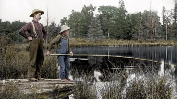 A man and a young boy stand on a wooden dock, fishing at a calm pond surrounded by grass and trees. The boy holds a long fishing rod, and both wear hats and old-fashioned clothes.