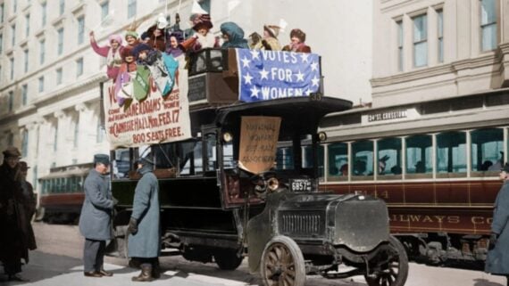 A group of women on a double-decker bus holds banners reading &ldquo;Votes for Women&rdquo; during a suffrage parade, while bystanders watch and streetcars are visible in the background.