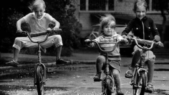 Three children ride bicycles through puddles, smiling and laughing. One child jumps with feet off the pedals. The scene is energetic, carefree, and joyful, with wind blowing their hair as they ride outdoors. The image is in black and white.