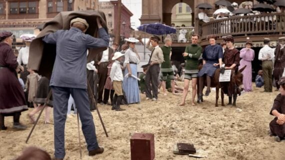 A vintage beach scene shows a photographer taking a photo of people in old-fashioned swimwear and clothes, with a crowd watching and a person riding a donkey holding a sign. The ground is sandy, and old buildings are visible.