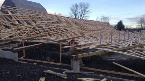 Collapsed wooden framework at a construction site, with a person in a hard hat inspecting the debris. Trees and a barn are visible in the background under a cloudy sky.