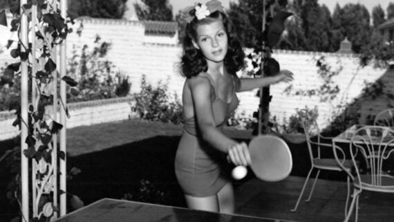 A woman in a swimsuit plays ping pong outdoors near a white brick wall and patio furniture, holding a paddle and ready to hit the ball.