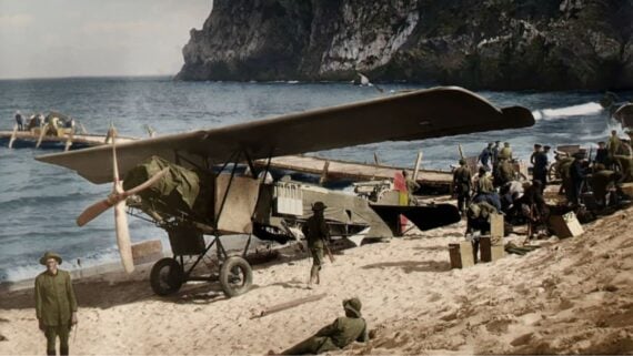 A vintage biplane sits on a sandy beach near the sea, surrounded by soldiers and equipment. Rocky cliffs rise in the background, and boats are visible on the water and shore.