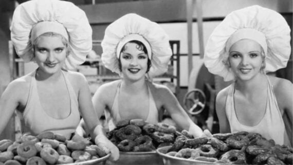 Three women wearing large chef hats and aprons smile while holding large trays filled with donuts in a bakery or kitchen setting. The image is in black and white.