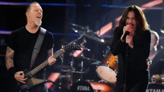 Two musicians perform on stage; one plays an electric guitar and sings, while the other holds a microphone and sings passionately. Drums and stage lights are visible in the background.
