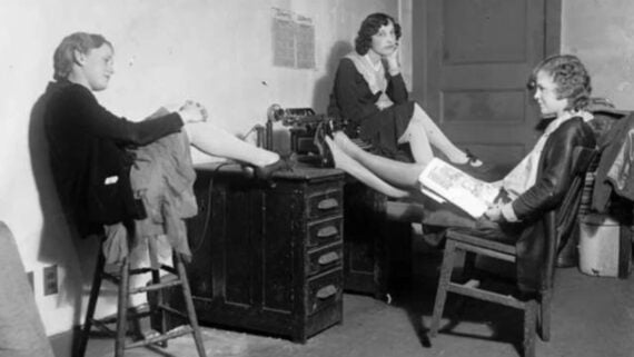Three women sit in a room, each with a leg propped up on a desk or chair. One holds a newspaper, another looks thoughtful, and a sewing machine is on the desk between them.
