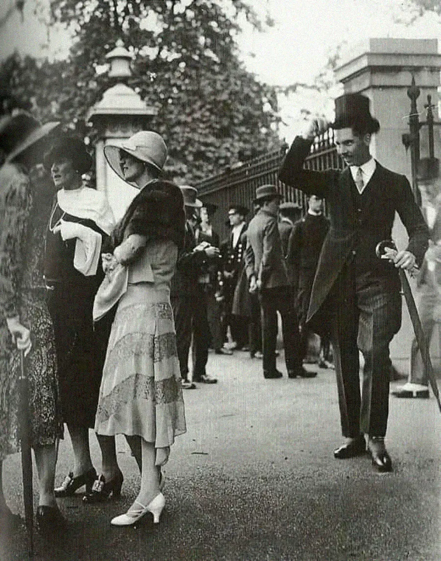 People in 1920s fashion stand and chat outside a gated park; women wear cloche hats and dresses, while a man in a suit and top hat lifts his hat in greeting. Others mill about in the background.