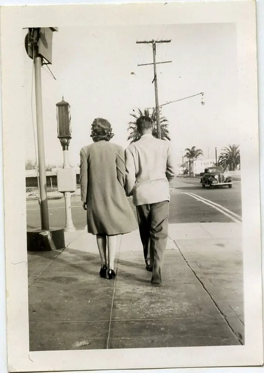 A woman and man walk side by side down a sidewalk in a vintage black-and-white photo. Palm trees, an old car, and a streetlamp are visible in the background.